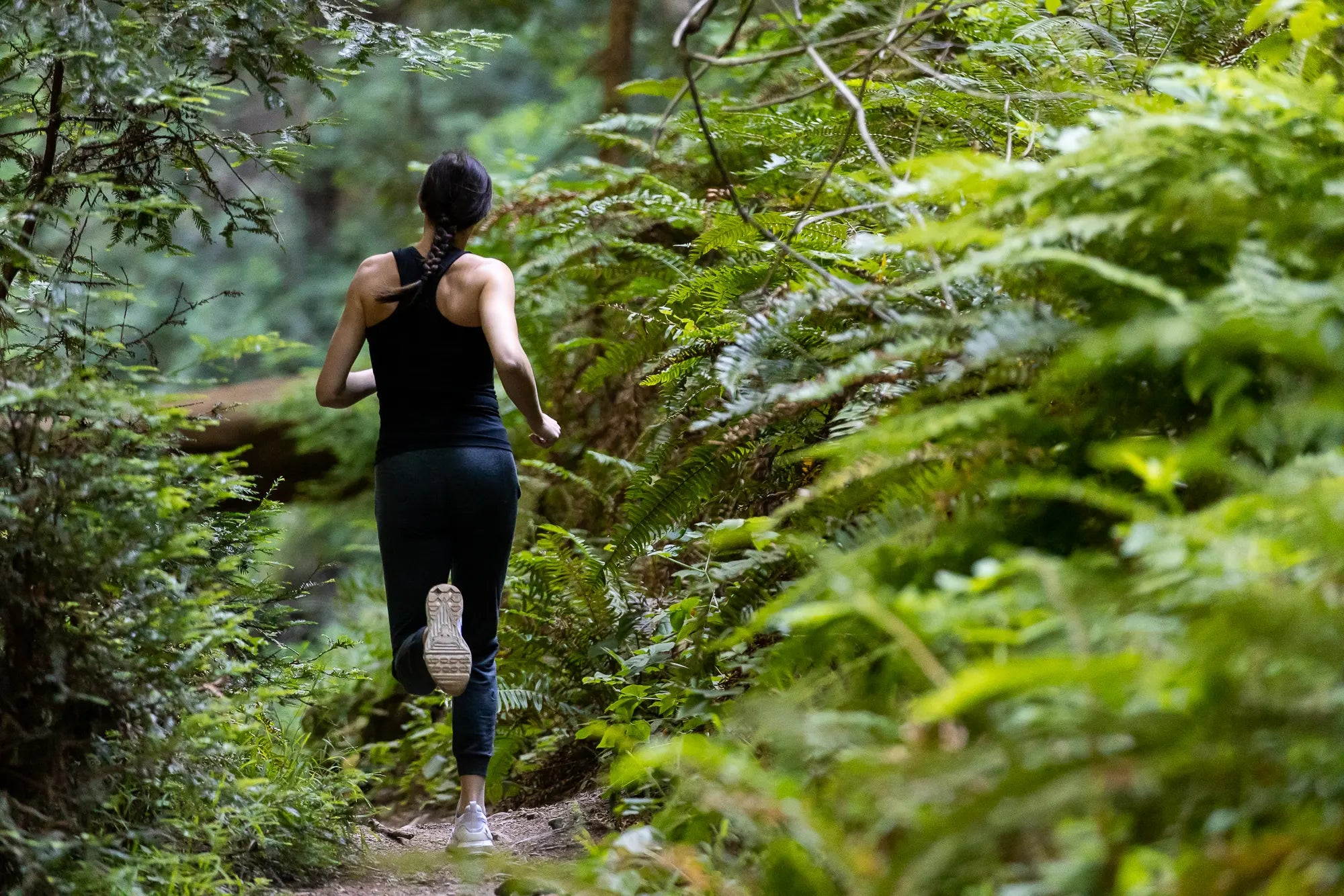 a woman wearing a Ridge Merino tank top and joggers goes for a trail run on a lush green trail