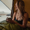 A woman smiles in a tent wearing the Merino wool bralette and drinking coffee