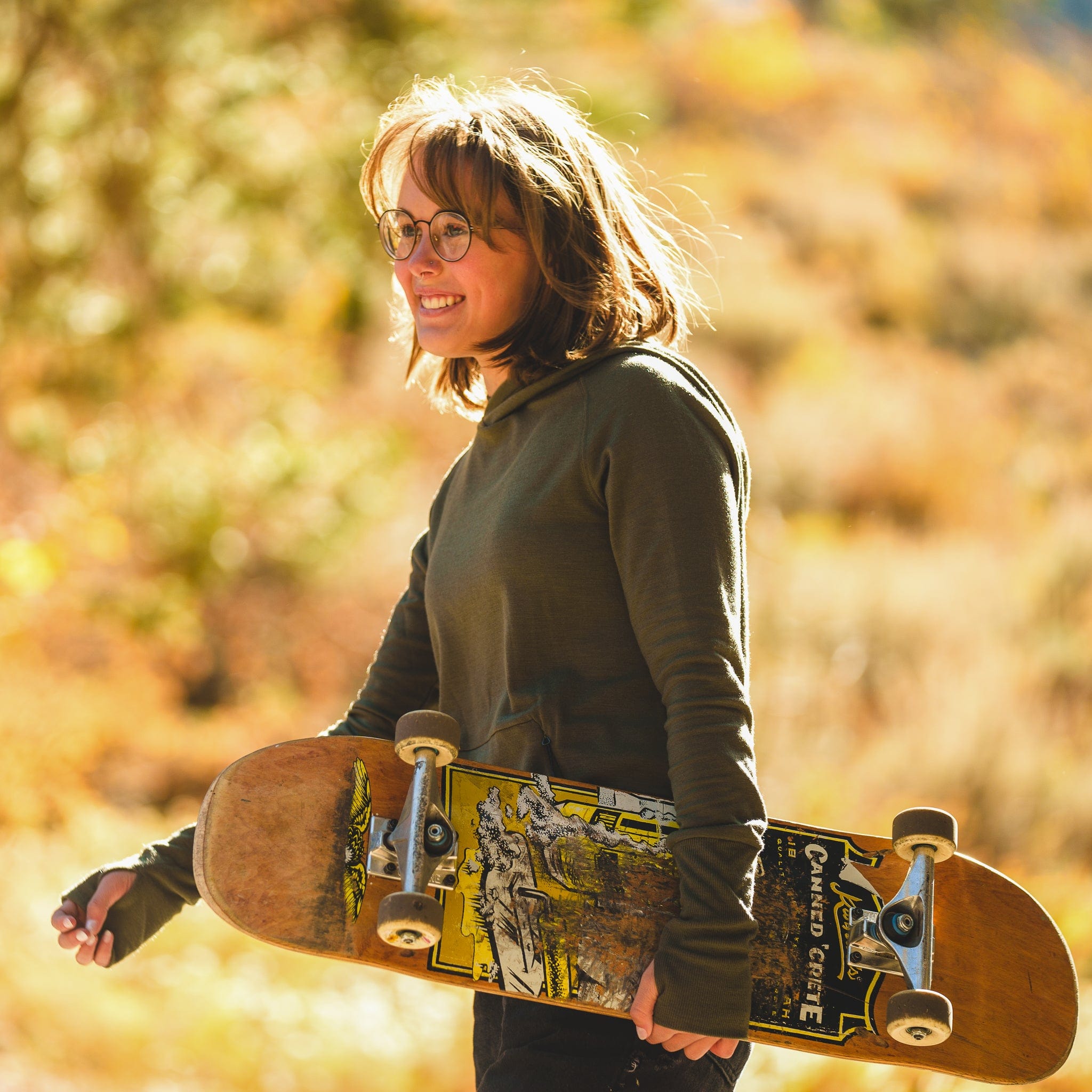 a woman carries a skateboard on a fall day wearing a Ridge Natural Hoodie