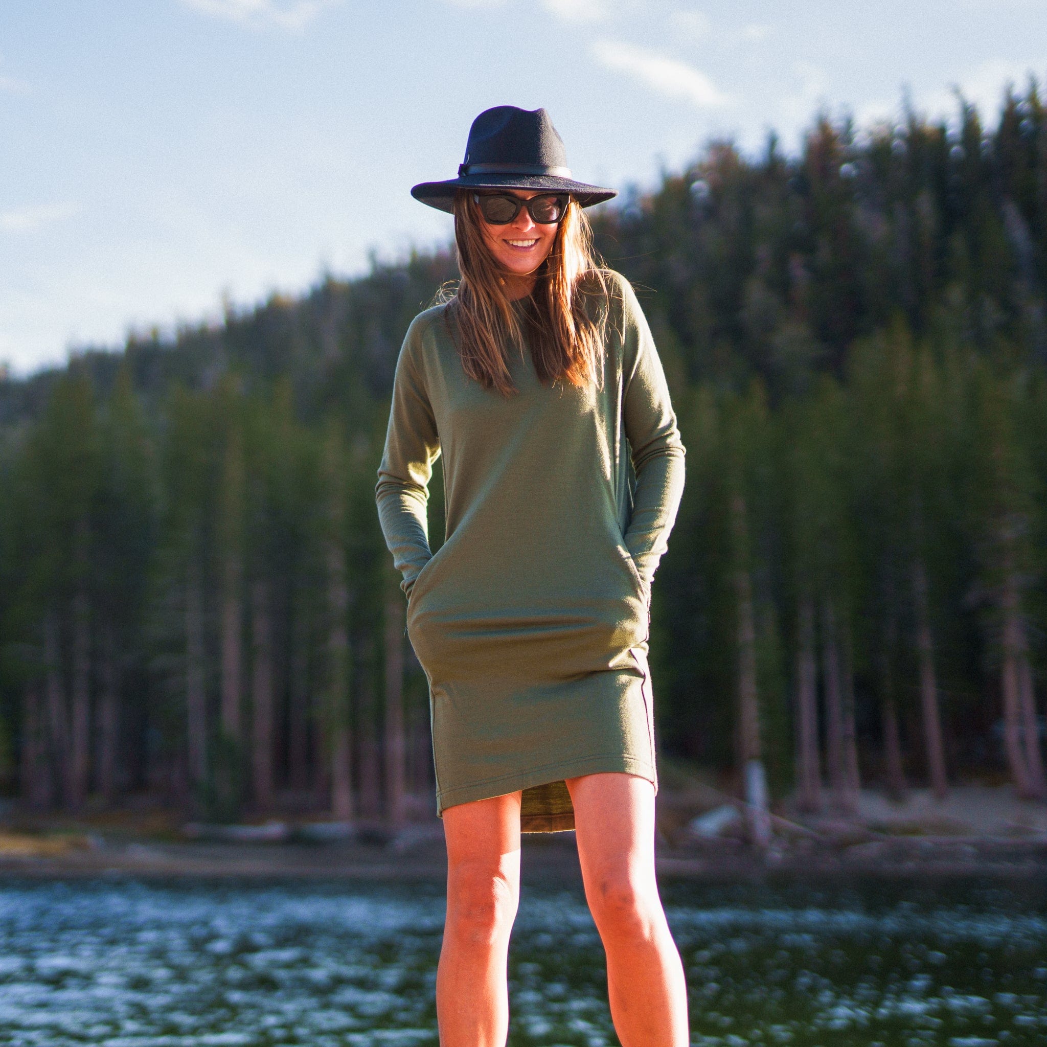 a woman stands by a lake wearing the Ridge Merino Hyde Dress and a hat and sunglasses