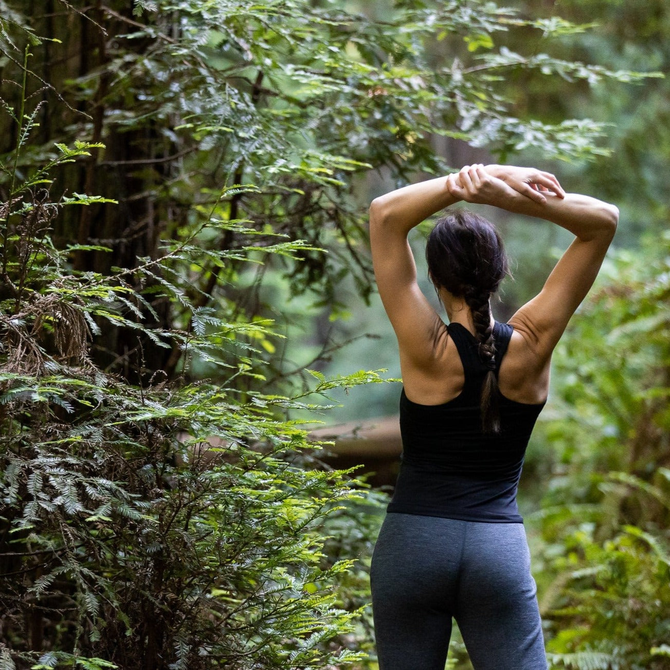 a woman stretching in a forest wearing a Ridge Merino racerback tank top